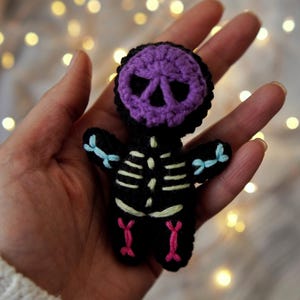 May include: A handmade crochet skeleton doll with a purple skull head and black body. The skeleton has light blue bows on the arms and pink accents on the legs. The background is blurred with bokeh lights.