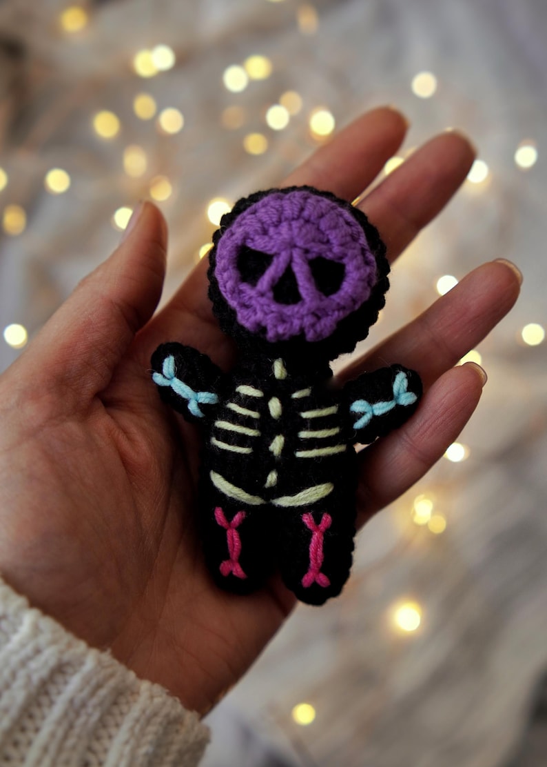 May include: A handmade crochet skeleton doll with a purple skull head and black body. The skeleton has light blue bows on the arms and pink accents on the legs. The background is blurred with bokeh lights.