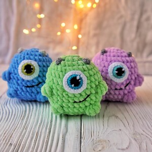 May include: Three crocheted plush toys shaped like monsters with one large eye each. The monsters are blue, green, and purple.