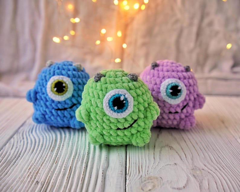May include: Three crocheted plush toys shaped like monsters with one large eye each. The monsters are blue, green, and purple.