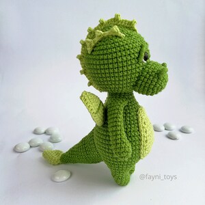 May include: A green crocheted dragon toy with a spiky green crest, a small wing, and a long tail. The dragon has a cute face with black eyes and a small nose. The toy is standing on a white surface with white pebbles scattered around it. The text '@fayni_toys' is visible in the bottom right corner of the image.