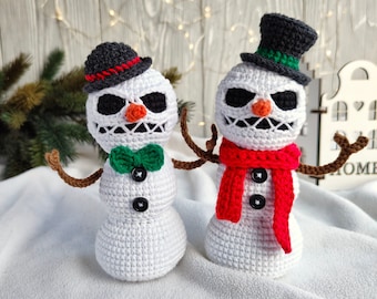 May include: Two crocheted snowmen with black hats and white bodies. One snowman wears a green bow tie and the other wears a red scarf. Both snowmen have black eyes and a stitched-on smile.