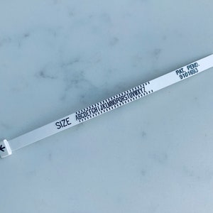 May include: White plastic ring sizer with black lettering. The sizer has a black arrow on the left side and the letters A through Z printed on the right side. The text "SIZE" is printed on the left side of the sizer. The text "PAT. PEND. 9101693" is printed on the right side of the sizer.