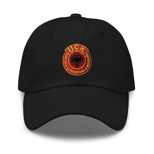 May include: Black baseball cap with an embroidered yellow circle containing the text "UCK USHTRIA CLIRIMTARE E KOSOVS" and a red Albanian double-headed eagle.