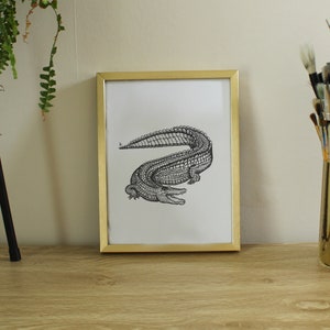 May include: A gold framed print of a black and white line drawing of an alligator. The print is sitting on a wooden surface with a gold container of paint brushes.