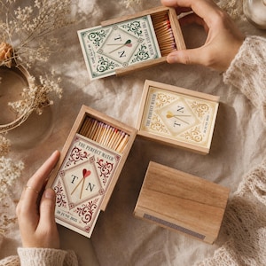 Peut inclure: Trois boîtes d'allumettes en bois avec des tiroirs coulissants contenant des allumettes. Chaque boîte présente une étiquette en losange avec le texte THE PERFECT MATCH, les lettres T et N, et la date 30 JUNE 2025 en vert, or et rouge.