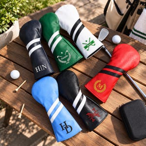 Könnte beinhalten: Ein Set aus sechs Golfschlägerhauben aus Leder in Marineblau, Grün, Weiß, Rot, Hellblau und Schwarz. Jede Haube hat zwei weiße oder schwarze Streifen und eine individuelle Bestickung, darunter Initialen wie H|N, HD, M sowie Namen wie Danielle und Batoul.