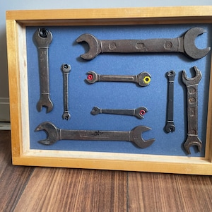 May include: A wooden shadow box frame containing a collection of seven vintage metal spanners mounted on a blue background. The spanners vary in size and design, with some featuring small red and yellow nuts attached to their open ends.