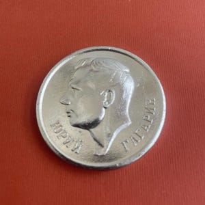 Puede incluir: Una moneda plateada con un perfil de hombre y texto cirílico alrededor del borde, sobre un fondo rojo. El diseño de la moneda es detallado, con los rasgos del hombre claramente visibles. La moneda es de forma circular.