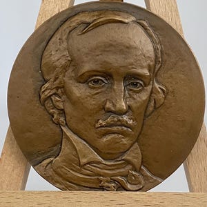 Puede incluir: Una escultura en relieve circular de color bronce que representa el busto de un hombre, con rasgos faciales detallados, incluyendo un bigote prominente y un cuello alto. La escultura se exhibe en un pequeño caballete de madera.