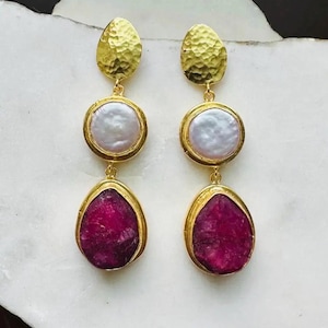 May include: A pair of gold-tone drop earrings featuring a hammered gold disc at the top, a round white pearl in the center, and a teardrop-shaped deep pink gemstone at the bottom, all set in gold-colored metal frames with a polished finish.