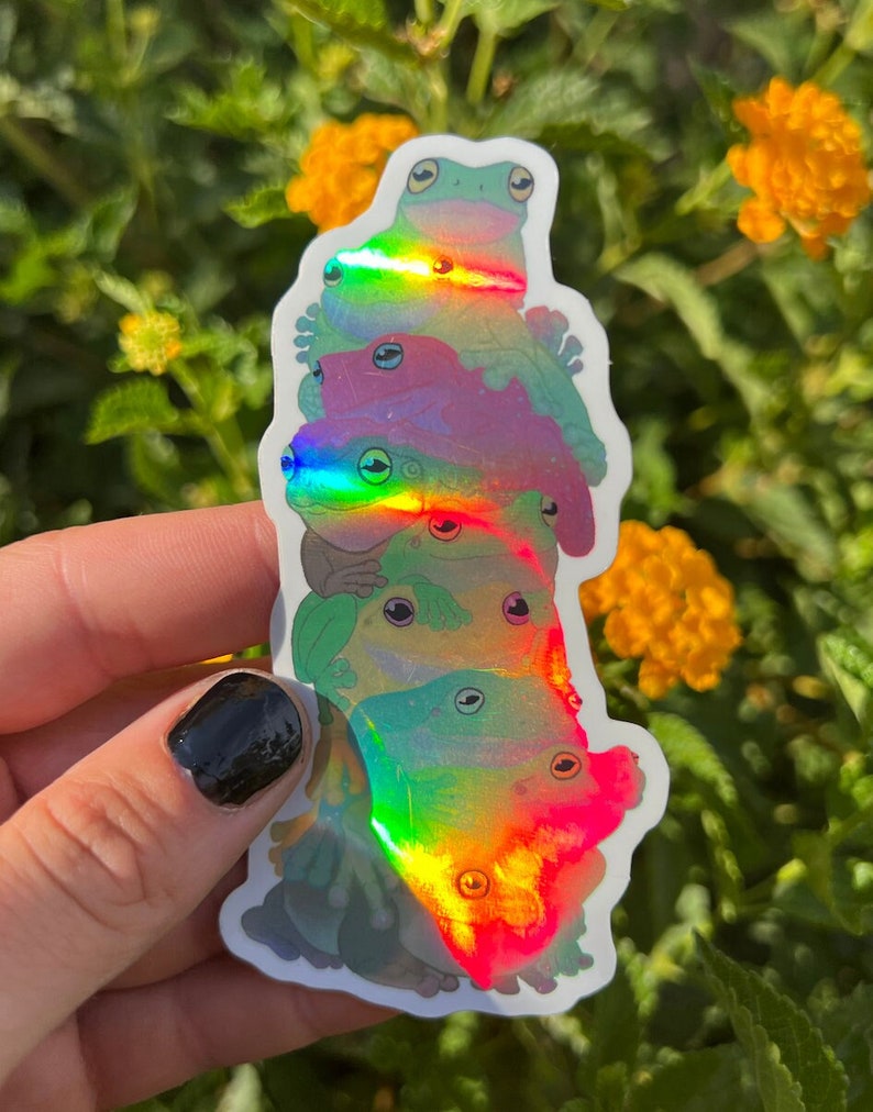 May include: A stack of iridescent frog stickers with a rainbow holographic finish. The frogs are stacked on top of each other, with the largest frog at the bottom and the smallest frog at the top.