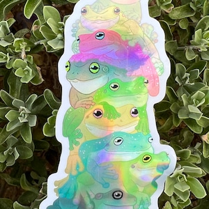 May include: A stack of six iridescent frog stickers with a rainbow effect. The frogs are stacked on top of each other, with the largest frog at the bottom and the smallest frog at the top. The frogs are all facing the same direction, with their eyes open and their mouths closed.