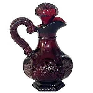 Può includere: Una brocca vintage in vetro rosso rubino con manico e tappo strutturato. La brocca presenta dettagli ornati, tra cui un manico strutturato e pannelli decorativi sul corpo. Il beccuccio è curvo e la base ha una forma scura e sfaccettata.