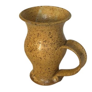 May include: A ceramic mug with a speckled tan and brown finish. The mug features a flared rim, a bulbous body that tapers toward the base, and a curved handle attached to the side. The surface has a glossy texture with dark brown speckles.