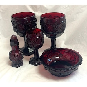 Op de afbeelding: Een verzameling robijnrood glaswerk, waaronder kelken, een klein wijnglas, een kom en een kleine fles. De stukken hebben een reliëf bloemen- en geometrisch patroon. De set wordt gepresenteerd op een wit oppervlak.