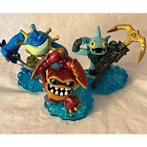 May include: Three colourful action figures on blue water-like bases. The figures include a blue and green creature with a trident, a red crab-like character, and a teal fish-like figure with a weapon. The toys are from the Skylanders series.