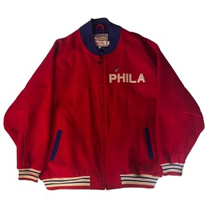 May include: A red zip-up bomber jacket featuring a blue collar, blue-trimmed pockets, and striped cuffs. The word PHILA is embroidered in white on the chest. The interior label displays the Hardwood Classics logo and Mitchell & Ness branding.