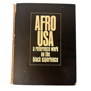 Puede incluir: Un libro negro con el título "AFRO USA" en letras doradas grandes. Debajo del título, el texto "a reference work on the black experience" en un rectángulo blanco. Los bordes del libro muestran desgaste.