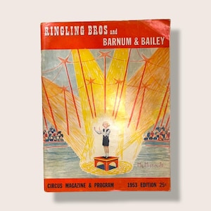 Peut inclure: Magazine et programme de cirque Ringling Bros. et Barnum & Bailey de 1953. La couverture présente un dessin d'un artiste sur une petite scène, éclairé par des projecteurs, avec un public en arrière-plan. Le magazine est dans les tons de rouge, jaune, bleu et blanc.
