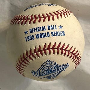 May include: A white Rawlings baseball with red stitching featuring blue text that reads OFFICIAL BALL 1995 WORLD SERIES and a 1995 World Series logo. The ball has a smooth leather surface and is designed for professional baseball play.