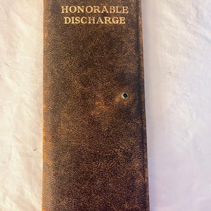 May include: A vintage, rectangular brown leather document holder with the words "HONORABLE DISCHARGE" in gold lettering. The cover shows signs of wear and a small metal eyelet. The overall aesthetic suggests a historical or military theme.