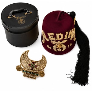 May include: A maroon felt fez hat with gold lettering reading NEDIM No. 19, a gold sword and crescent emblem, and a long black tassel. It is accompanied by a black cylindrical carrying case and a gold crescent-shaped brooch with rhinestones.