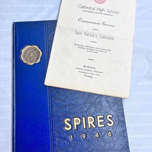 Op de afbeelding: Een blauw hardcover jaarboek met gouden letters met de tekst "SPIRES 1946" wordt getoond met een crèmekleurig afstudeerprogramma. Het jaarboek heeft een gouden zegel en het programma is van Cathedral High School.