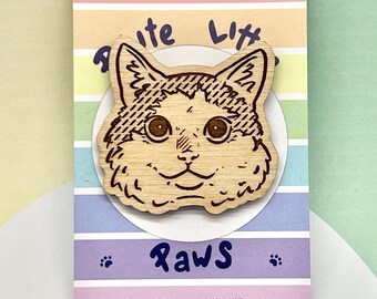 May include: A wooden cat-shaped pin featuring a laser-engraved feline face with detailed fur and whiskers. The pin is mounted on a rectangular card with horizontal pastel stripes and the text Polite Little Paws printed in dark blue.