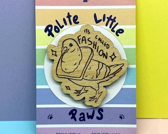 May include: A wooden pin featuring a pigeon wearing a slice of bread around its neck, with the text "POLITE LITTLE PAWS" and "ITS CALLED FASHION" engraved on the surface, mounted on a rectangular card with horizontal pastel stripes.