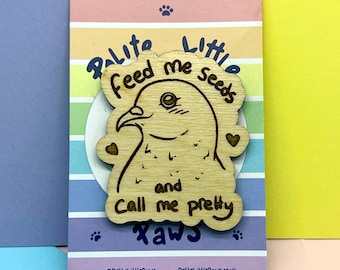 May include: A light-colored wooden pin shaped like a bird, featuring the engraved text "Feed me seeds and Call me pretty" with two small hearts. The pin is mounted on a rectangular backing card with horizontal rainbow stripes and paw print icons.