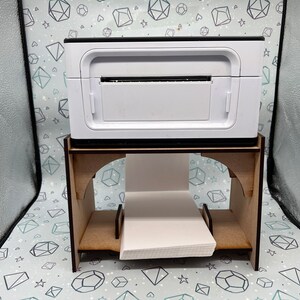 May include: A white label printer sits on a wooden stand with a stack of white labels underneath. The printer has a black top and a black base.