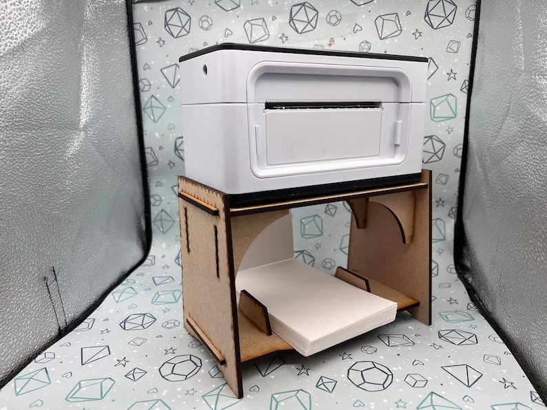 May include: A white photo printer sits on a wooden stand with a stack of photo paper underneath. The stand has a cutout in the back to allow for cords to be plugged in.