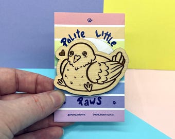 May include: A light-colored wooden pin featuring a laser-engraved illustration of a round bird. The pin is attached to a rectangular card with horizontal pastel stripes and the text Polite Little Paws printed in dark blue ink.