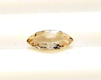 Puede incluir: Una gema de talla marquesa, de color melocotón claro. La gema está facetada, con un extremo puntiagudo en cada lado. La piedra está sobre un fondo blanco.
