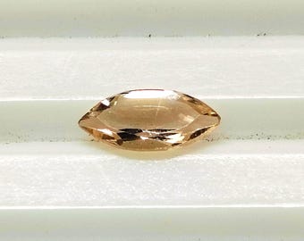 Puede incluir: Una gema facetada, de talla marquesa, con un tono cálido color melocotón. La gema se presenta sobre un fondo blanco, mostrando sus superficies pulidas y su claridad interna. La forma alargada y la refracción de la luz de la gema son claramente visibles.