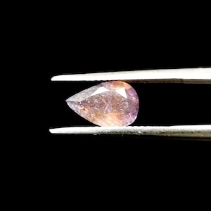 May include: A pear-shaped gemstone held by tweezers against a black backdrop. The gem displays a blend of colours, including purple, orange, and brown. The stone's facets reflect light, producing a sparkling effect. The overall impression is one of elegance and luxury.