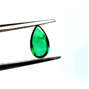 May include: A pear-shaped emerald gemstone, held by tweezers, with a deep green colour and a faceted cut.