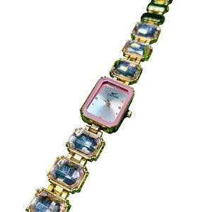 Puede incluir: Reloj dorado con esfera rectangular rosa y correa adornada con grandes cristales rectangulares transparentes. La esfera del reloj tiene la palabra "CRETON" impresa. La correa es de metal dorado.