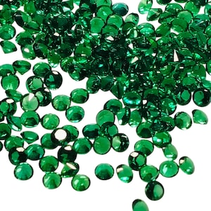 Puede incluir: Un conjunto de pequeñas piedras preciosas verde esmeralda facetadas. Las gemas, de forma redonda u ovalada, están talladas para reflejar la luz. Se dispersan sobre un fondo blanco, creando un efecto brillante.