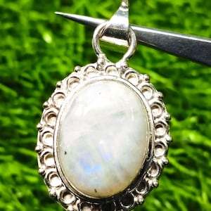 Op de afbeelding: Ovale zilveren hanger met een grote, iriserende, witte edelsteen, gezet in een filigraam frame. De hanger wordt door een pincet vastgehouden tegen een vage groene achtergrond, waarbij de details van de sieraden worden benadrukt.
