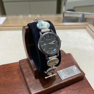 Op de afbeelding: Een zilverkleurig quartz horloge met een zwarte wijzerplaat en het woord "Forever" erop. De horlogeband heeft abalone schelp accenten. Het horloge wordt getoond op een houten standaard met een zwart fluwelen kussen.