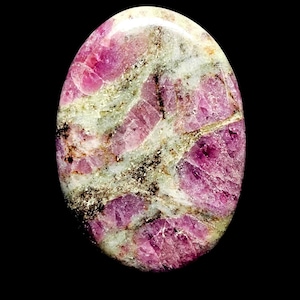 May include: Oval-shaped gemstone with a polished surface. The stone features a mix of pink, white, and gray hues, with darker pink and black inclusions. The gemstone is set against a black background.