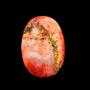 Puede incluir: Una piedra preciosa pulida de forma ovalada con un color base rosa salmón y rojo coral vibrante. La superficie muestra una prominente veta diagonal irregular de inclusiones minerales de color marrón oscuro y oro metálico.