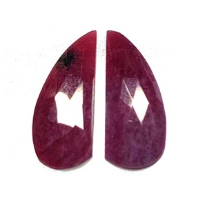 Może przedstawiać: Dwa fasetowane kamienie szlachetne w kształcie łezki. Kamienie mają głęboki, bogaty czerwony kolor z lekką zmiennością odcienia. Fasetki odbijają światło, tworząc efekt iskrzenia. Kamienie szlachetne są na białym tle.