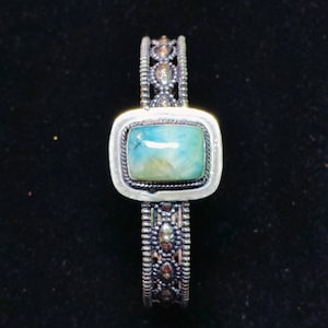 May include: A silver-toned bracelet with an oblong, light blue and yellow stone at the center. The band features intricate detailing with small, decorative elements. The bracelet is set against a dark background.