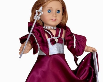 Puede incluir: Una muñeca con un vestido burdeos con detalles plateados y una corona plateada. El vestido tiene un ribete blanco y lentejuelas plateadas. La muñeca sostiene una varita plateada con una estrella en la parte superior.