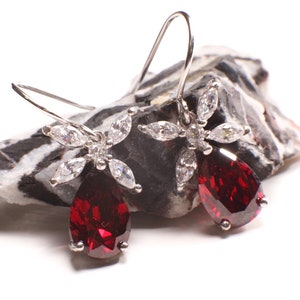 May include: A pair of silver dangle earrings with a red teardrop-shaped gemstone and a flower-shaped design with clear cubic zirconia stones.