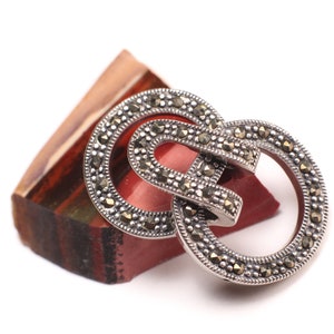May include: A silver brooch with a double circle design, set with marcasite stones. The brooch is resting on a piece of red and brown agate.