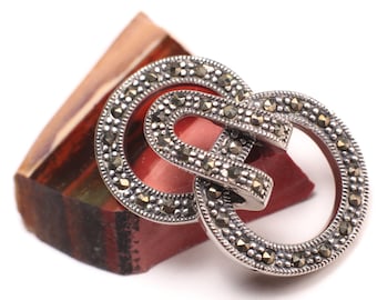 May include: A silver brooch with a double circle design, set with marcasite stones. The brooch is resting on a piece of red and brown agate.